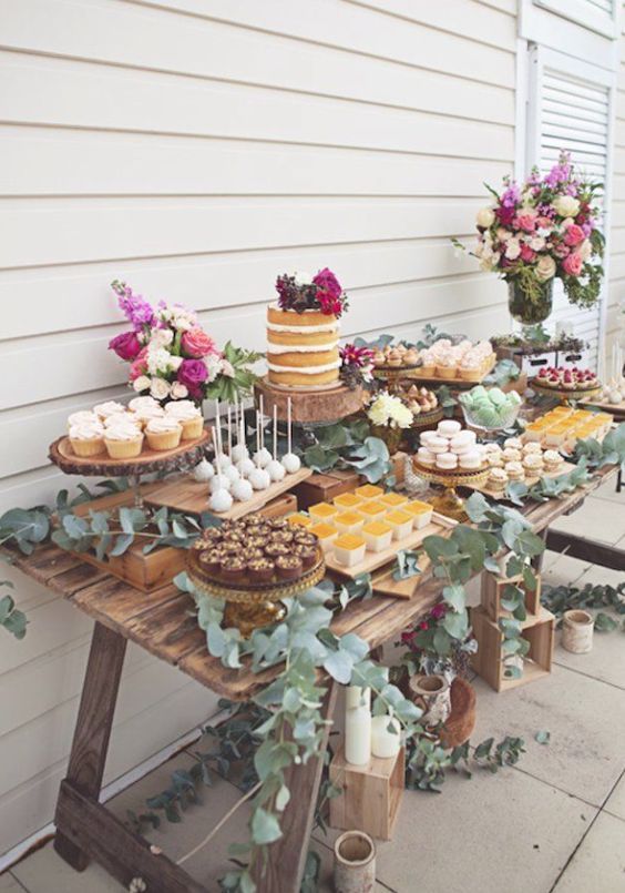 a dessert table decorated with eucalyptus features lots of desserts and sweets plus a cake