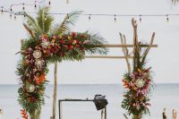 a driftwood tropical wedding arch decorated with greenery, with red, yellow, blush and orange blooms