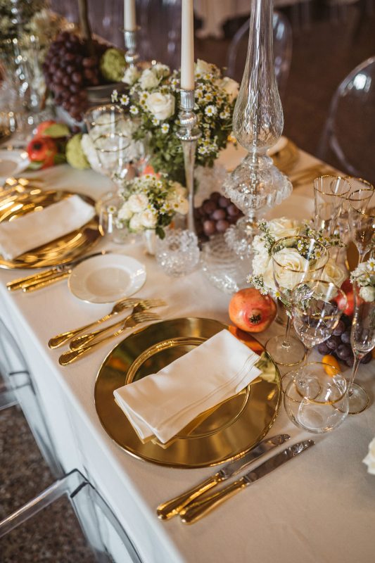 a jaw-dropping wedding tablescape with gold chargers and cutlery, candles in tall candleholders, glass and neutral blooms and greenery