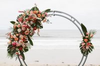 a round tropical wedding arch decorated with tropical leaves, orange, pink and white blooms