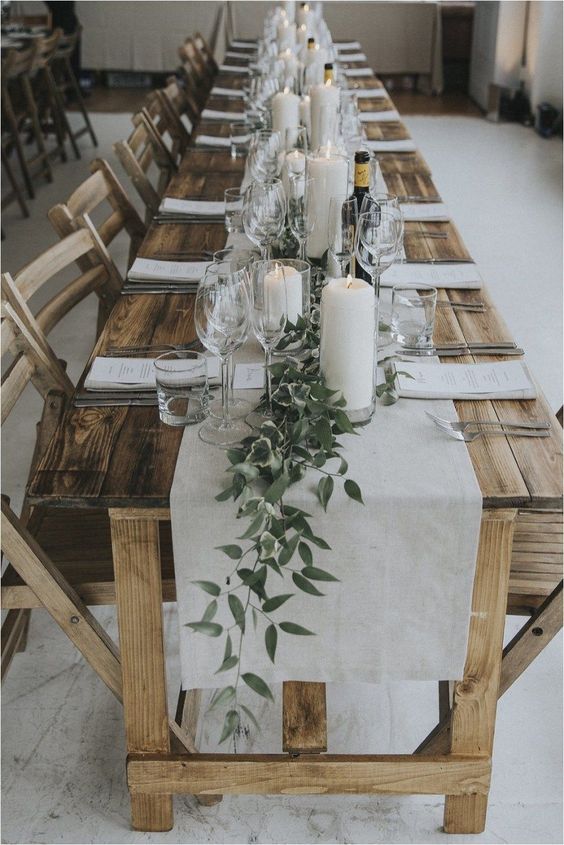 a rustic uncovered table with a white runner and greenery plus large white candles for a rustic rehearsal