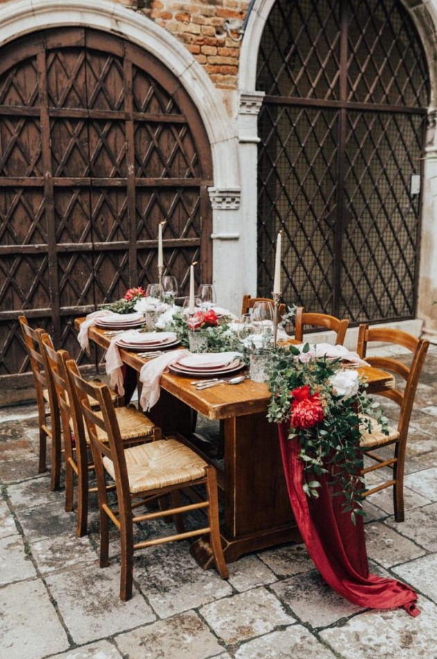 a sophisticated Venice wedding tablescape with a deep red table runner with white and red peonies and greenery, candles and pink plates