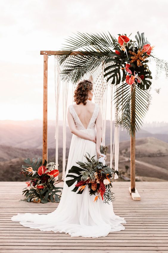 a statement wedding arch decorated with macrame, tropical leaves and red flowers is very bold
