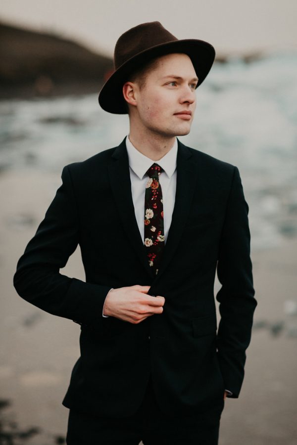 a stylish groom's look with a black suit, a white shirt and a dark floral tie plus a brown hat is lovely for an Icelandic wedding