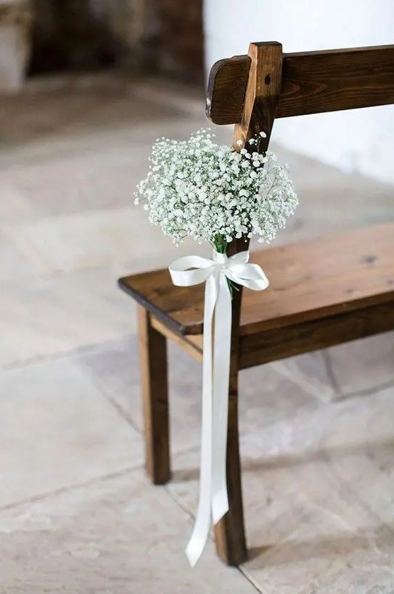 a wooden bench in the wedding aisle accented with a white silk bow with a baby's breath arrangement looks amazingly chic