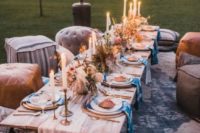 a boho chic picnic setting with candles, pampas grass, greenery and pink blooms, dip dyed napkins and leather ottomans