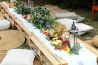 a boho picnic with a low table, with greenery, candle lanterns, neutral pillows and a vegetable and fruit table runner