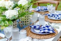 a bright and cool bbq rehearsal dinner tablescape with white hydrangeas and greenery, candleholders, woven placemats and plaid napkins