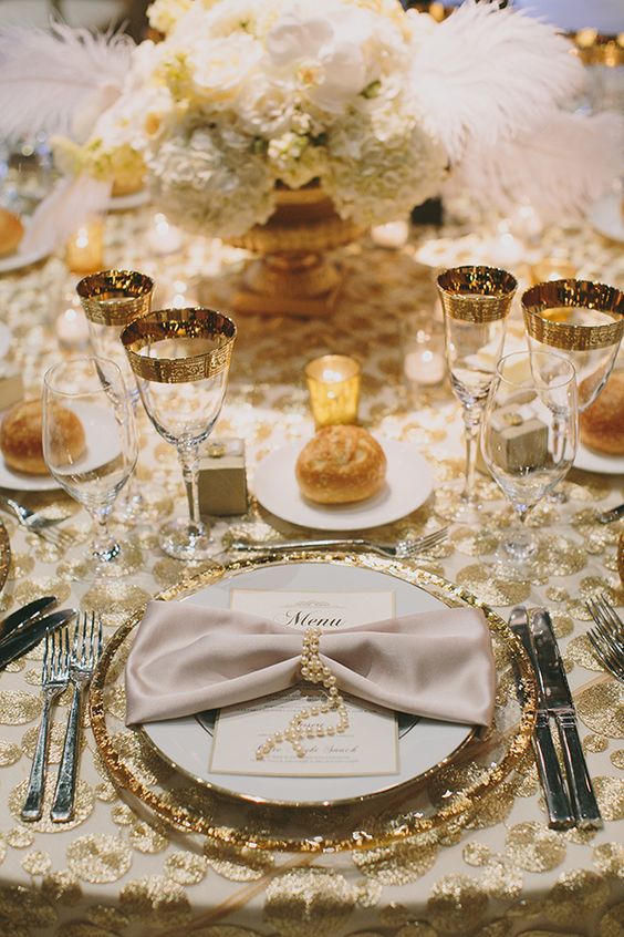 a chic 1920s tablescape with a white flower and feather centerpiece, gold rim glasses, candles and buns, gold rim chargers and pearls that tie the napkins