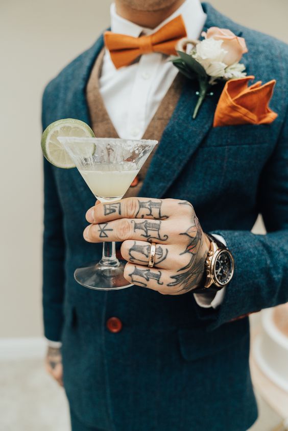 a colorful groom's look with a navy tweed suit, a beige waistcoat, a white shirt, an orange bow tie and a handkerchief and some hand tattoos