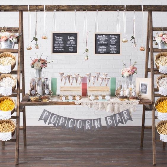a creative popcorn bar with a couple of ladders and a door, popcorn in wooden baskets, chalkboard signs and paper cones and toppings on the door