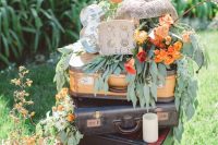 a creative wedding decoration of a stack of suitcases, greenery and bright blooms, a marquee light and a lantern with bold blooms
