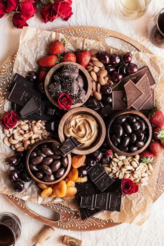 a decadent wedding chocolate board with various types of chocolate and candies, nuts, dried fruit, berries and red blooms around