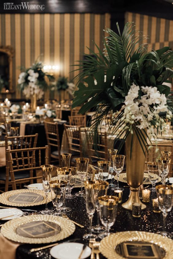 a glam and chic art deco tablescape with a black sequin tablecloth, gold placemats, a white bloom and greenery centerpiece and gold rim glasses