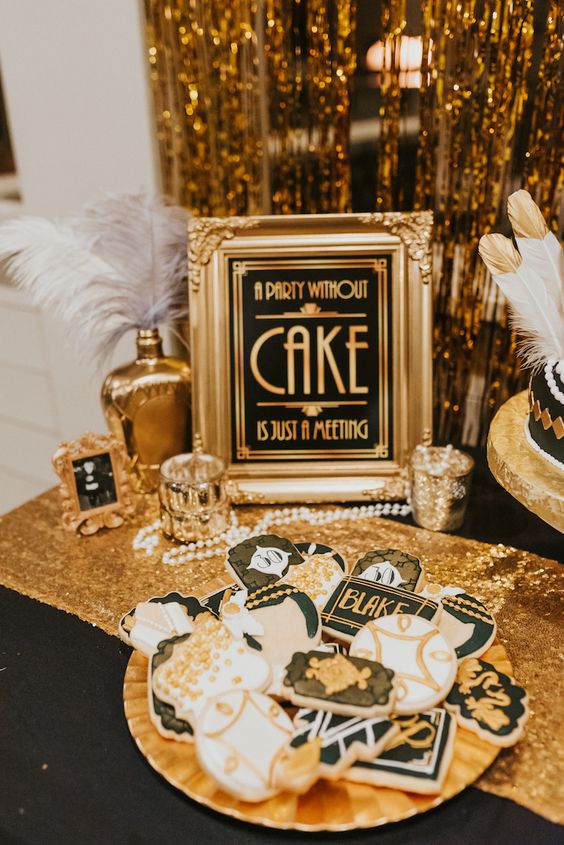 a glam gold and black dessert table with feathers, candles, pearls, sparkles, a sign and some themed cookies on a gold tray