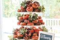 a large clear stand with lots of glazed donuts, and succulents and greenery plus a sign is your cool dessert table