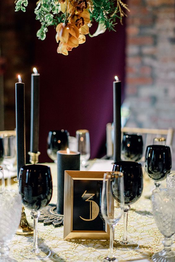 a lovely roaring 20s tablescape with a gold tablecloth, black candles and glasses, a black and gold sign and some greenery over the table