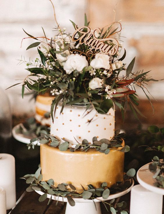 a lovely wedding cake with a gold tier, a white and gold polka dot tier, greenery, white blooms and a calligraphy topper is amazing