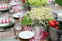 a rustic bbq rehearsal tablescape with a plaid runner and napkins, simple blooms in pots and fresh veggies and fruits in bowls