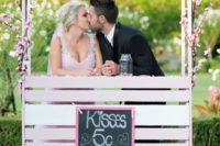 a spring-like kissing booth of white wood, with pink and white cherry blossom and with a chalkboard sign