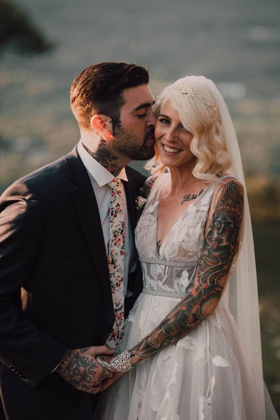 a stylish groom's look with a black pantsuit, a white shirt and a floral print tie, groom's tattoos on this hands and neck are shown in a great way