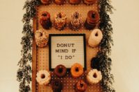 a stylish pegboard wall with holders and glazed donuts, with a sign and greenery and bloom garland on top
