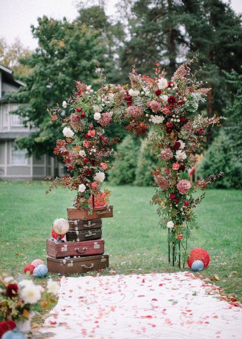a super lush and bright wedding arch with mauve, burgundy, white blooms and greenery and a stack of suitcases on one side is amazing