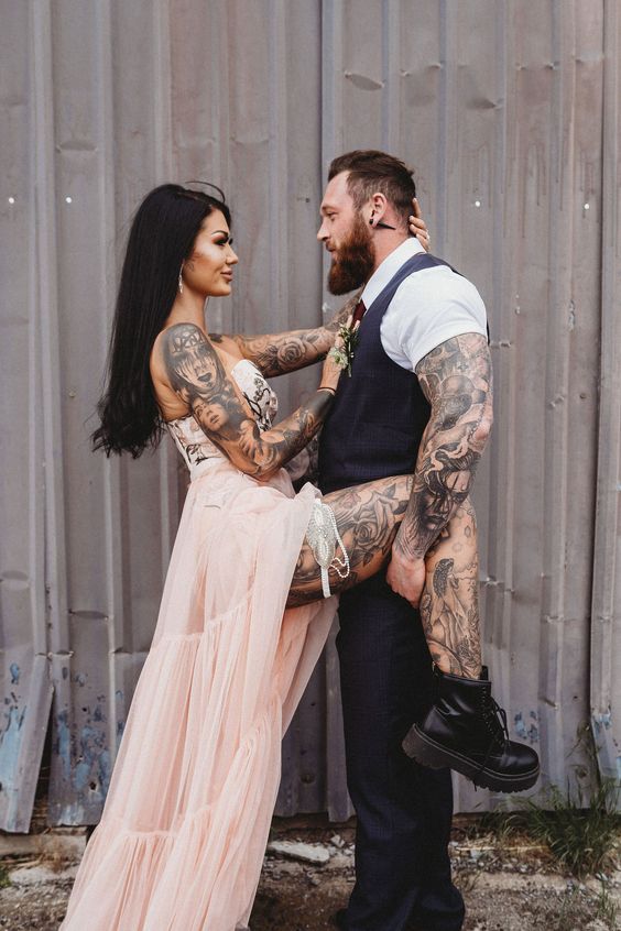 a tattooed couple, the groom wearing a grey waistcoat and pants, a white shirt with short sleeves to show off his arm tattoos