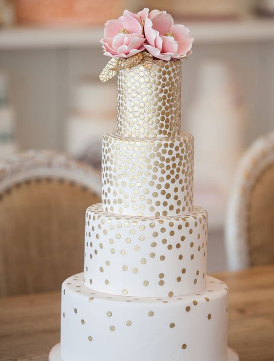 a white wedding cake with gold polka dots all over the cake and pink sugar blooms and gold leaves on top for a refined wedding