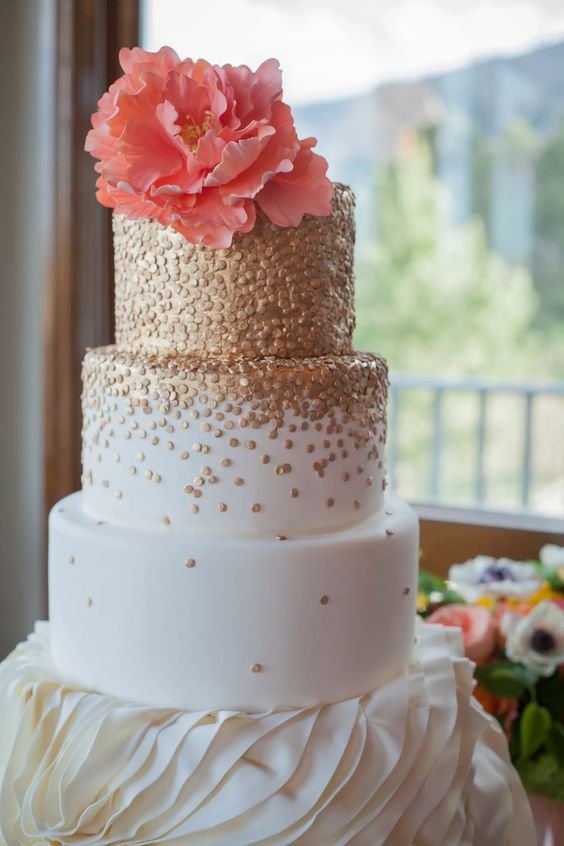 a white wedding cake with gold polka dots covering the top and the second tier, a coral sugar bloom on top for a bright wedding