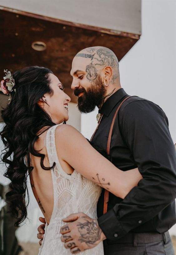 an informal groom's look with a black shirt and grey pants, amber leather suspenders with shown off head and hand tattoos