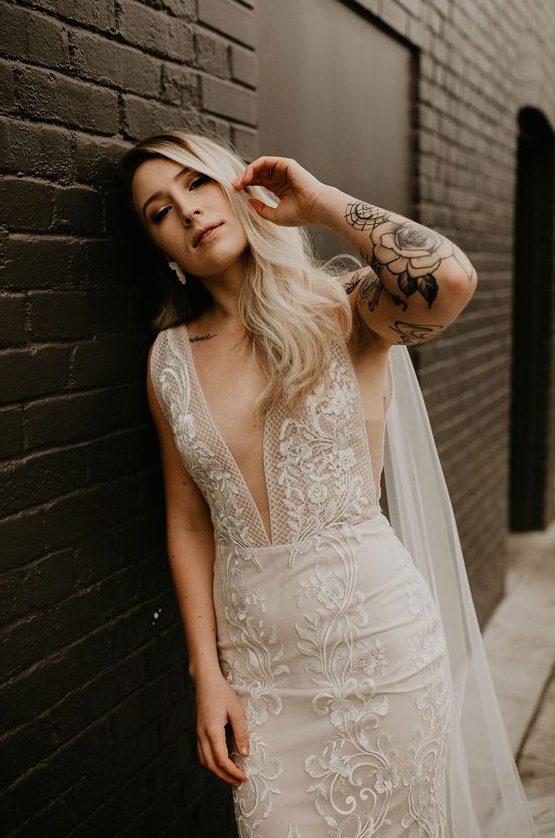 a sheath lace wedding dress with no sleeves and a plunging neckline plus tattoos that are shown off for a bold and sexy look