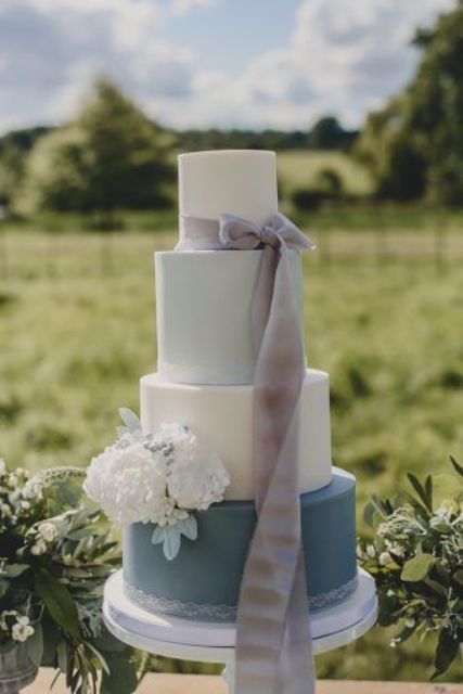 a beautiful white, light blue and navy wedding cake with lace detailing, a grey ribbon bow and white blooms for a spring wedding