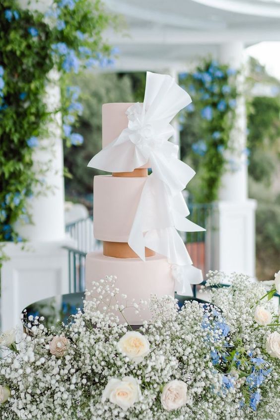 a blush three-tier wedding cake with white sugar bows is a stylish and refined idea for a modern wedding in neutrals and pastels