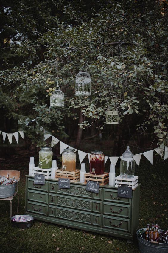 a chic and relaxed wedding drink bar of a green sideboard, crates with lemonade, cages on the trees and buntings is amazing