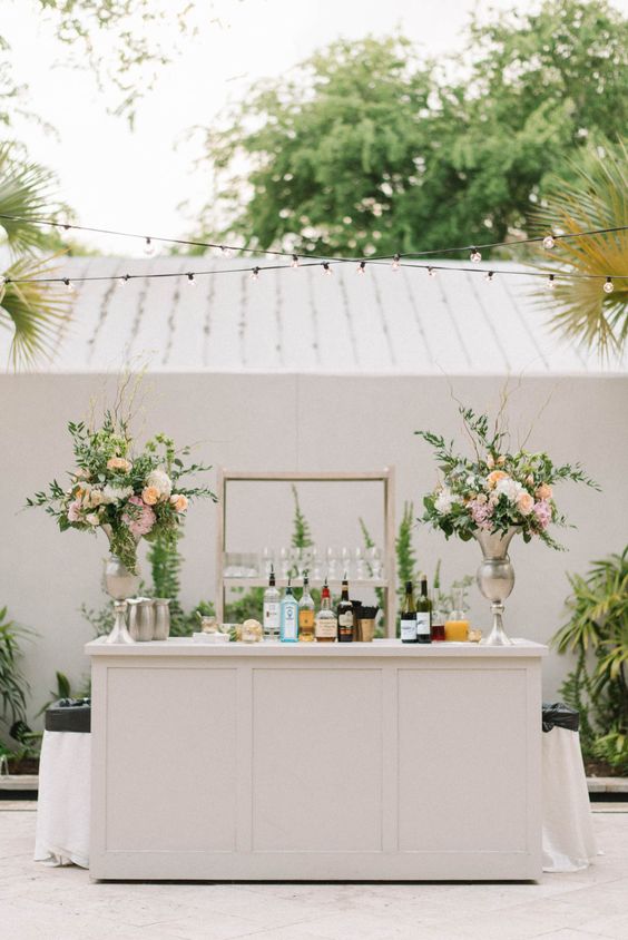 a chic wedding bar of a white bar stand, greenery and pastel floral arrangements, an open shelf is a lovely idea