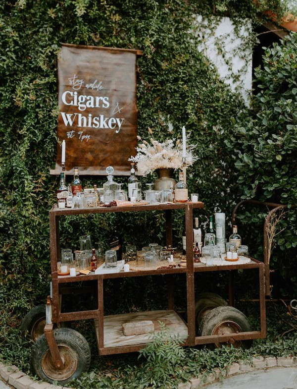 a cigar and whiskey bar made of a vintage farm trolley and a leather sign, decorated with neutral blooms and grasses and candles