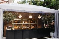 a cool glam wedding drink bar with a black bar stand, with gilded open shelves, potted trees and bubble glass pendant lamps