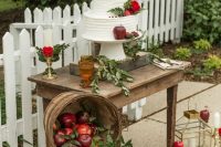 a delicate wedding dessert table with greenery and red blooms, a wooden basket with apples and greenery, a white wedding cake decorated wtih red apples and blooms