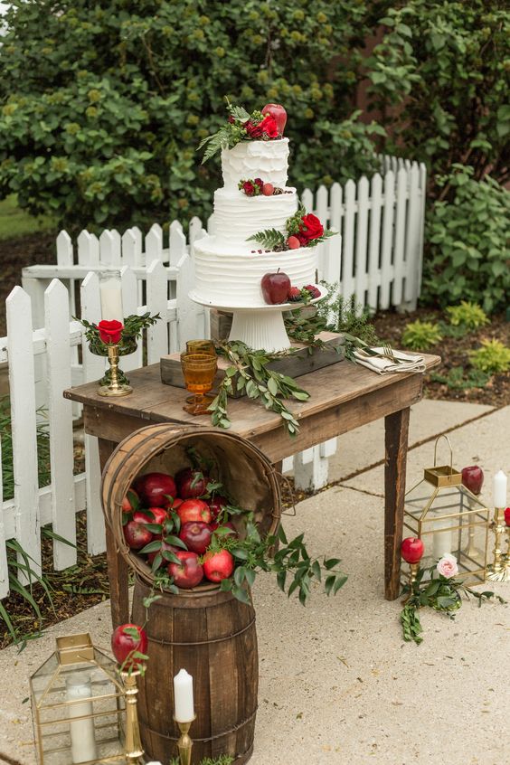 a delicate wedding dessert table with greenery and red blooms, a wooden basket with apples and greenery, a white wedding cake decorated wtih red apples and blooms