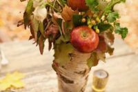 a fall wedding centerpiece of a vase wrapped with bark, various foliage, berries and apples plus some pinecones for a rustic or woodland wedding