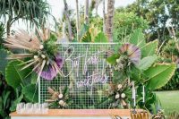 a fantastic tropical glam wedding bar of a white planked stand, fronds, blooms and tropical leaves and a neon calligraphy sign is chic