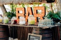 a gorgeous tropical wedding bar with a roof, a stand of stained wood and barrels, a marquee sign and tropical leaves is amazing