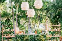 a gorgeous tropical wedding drink bar of a stand covered with fall leaves and wihte blooms, with pink open shelving units and woven pendant lamps