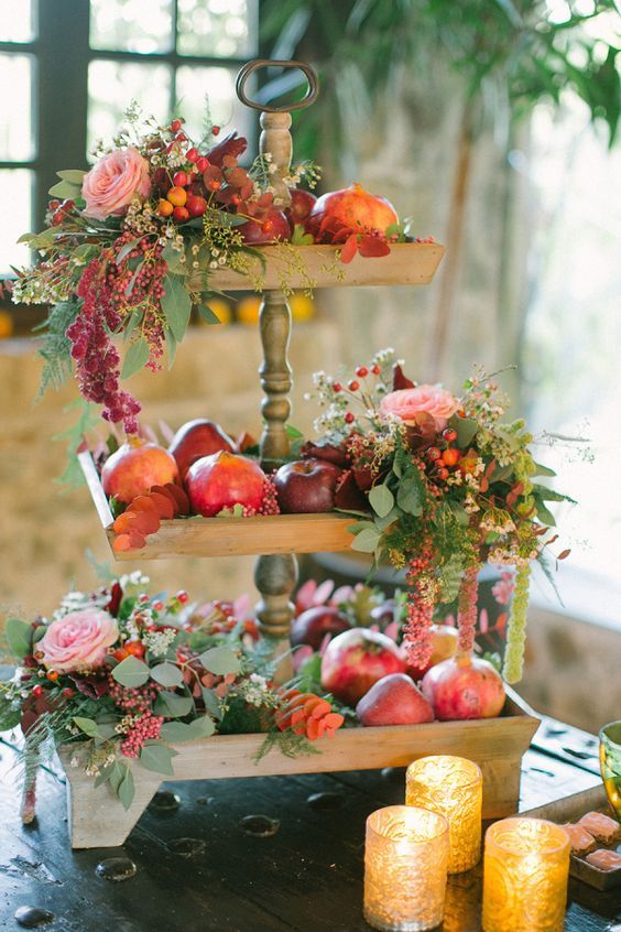 a lovely rustic fall wedding decoration of a tiered wooden stand with apples, pomegranates, pink blooms, berries and greenery