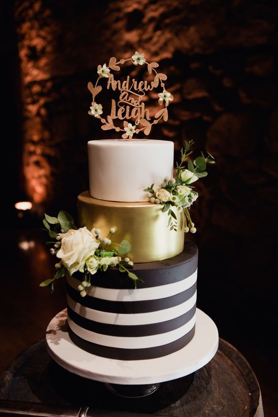 a modern wedding cake with a black and white striped tier, a gold one and a white one, with white blooms and a calligraphy topper