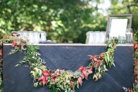 a pretty and chic wedding drink bar of a stand clad with black chevron planks and with a greenery and bodl leaf garland is a lovely idea