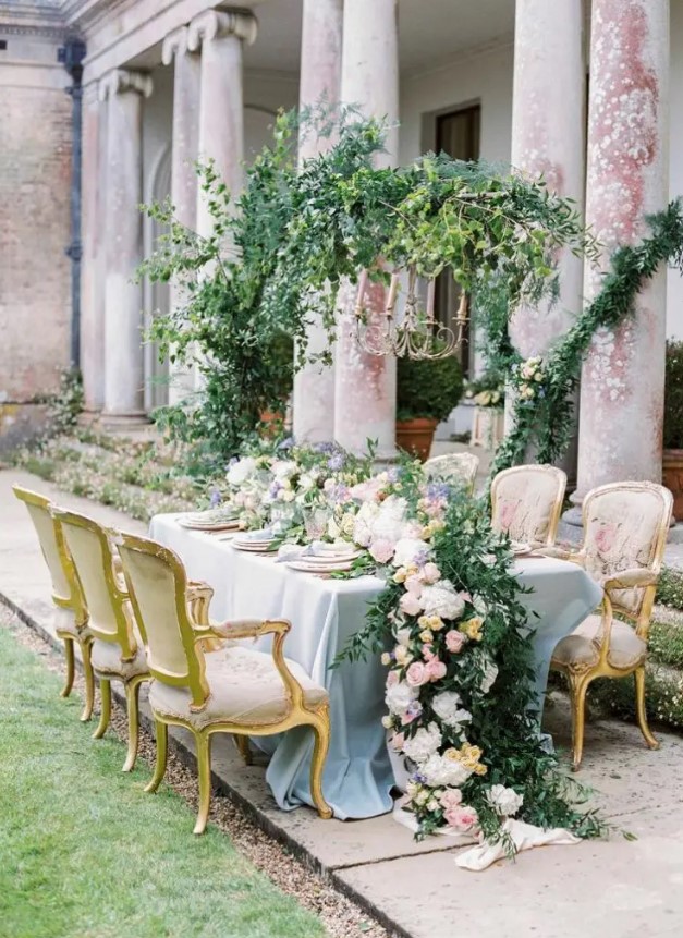 a refined garden wedding tablescape with a lush greenery and pastel bloom runner, pastel plates and linens and refined chairs