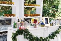 a refined wedding drink bar of a stand and a large open shelving unit, a greenery garland and lots of glasses and bright blooms in a vase is amazing
