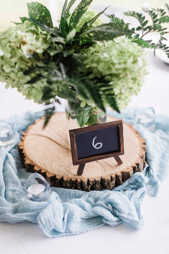 a rustic or woodland wedding centerpiece of a wood slice with a chalkboard table number and a greenery arrangement and a dusty blue table runner